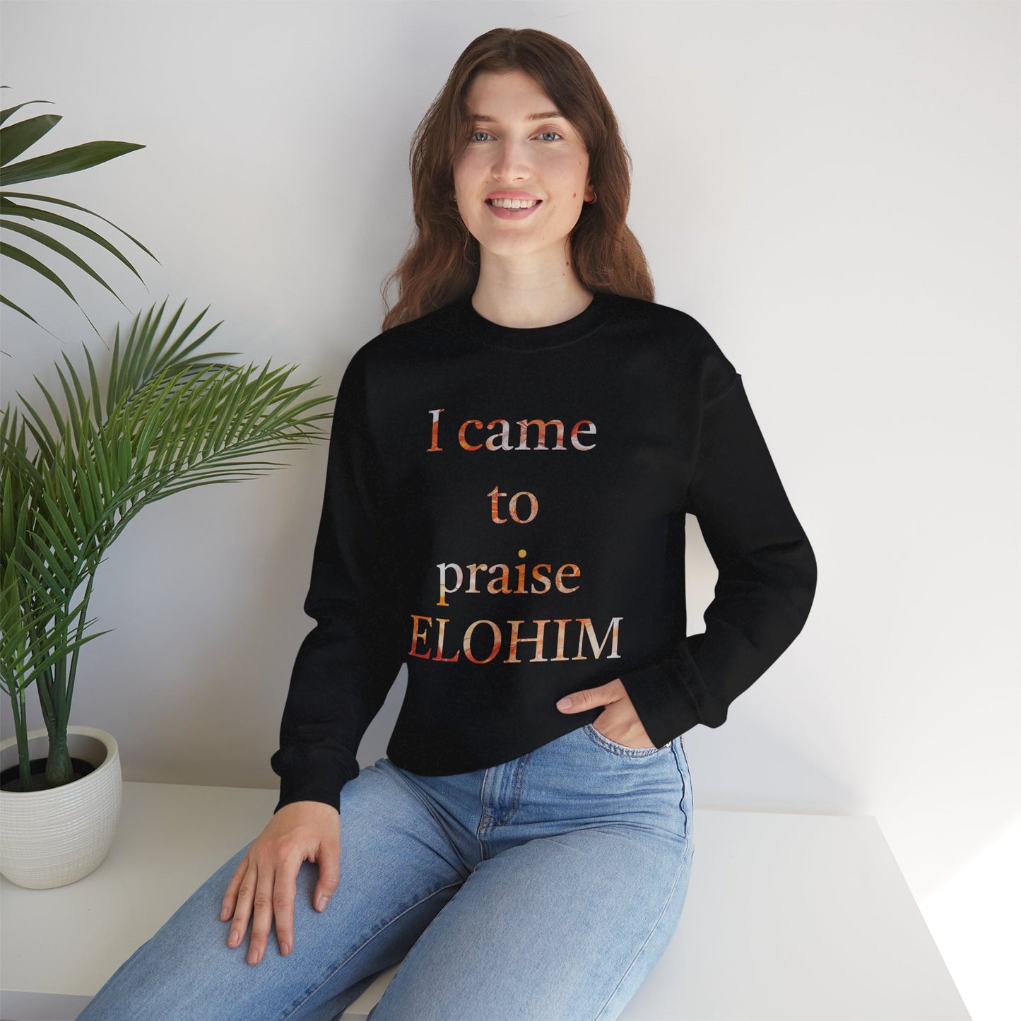 A woman smiling and wearing a black crewneck sweatshirt with the text 'I came to praise ELOHIM' in stylized lettering.