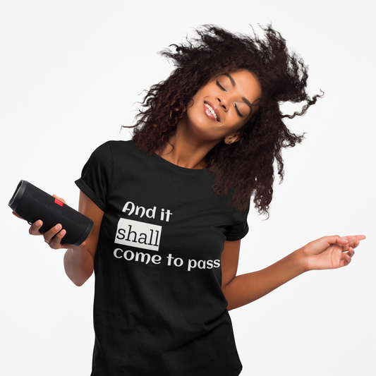 A woman with curly hair is smiling while wearing a black crew neck t-shirt with the phrase 'And it shall come to pass' printed on the front.