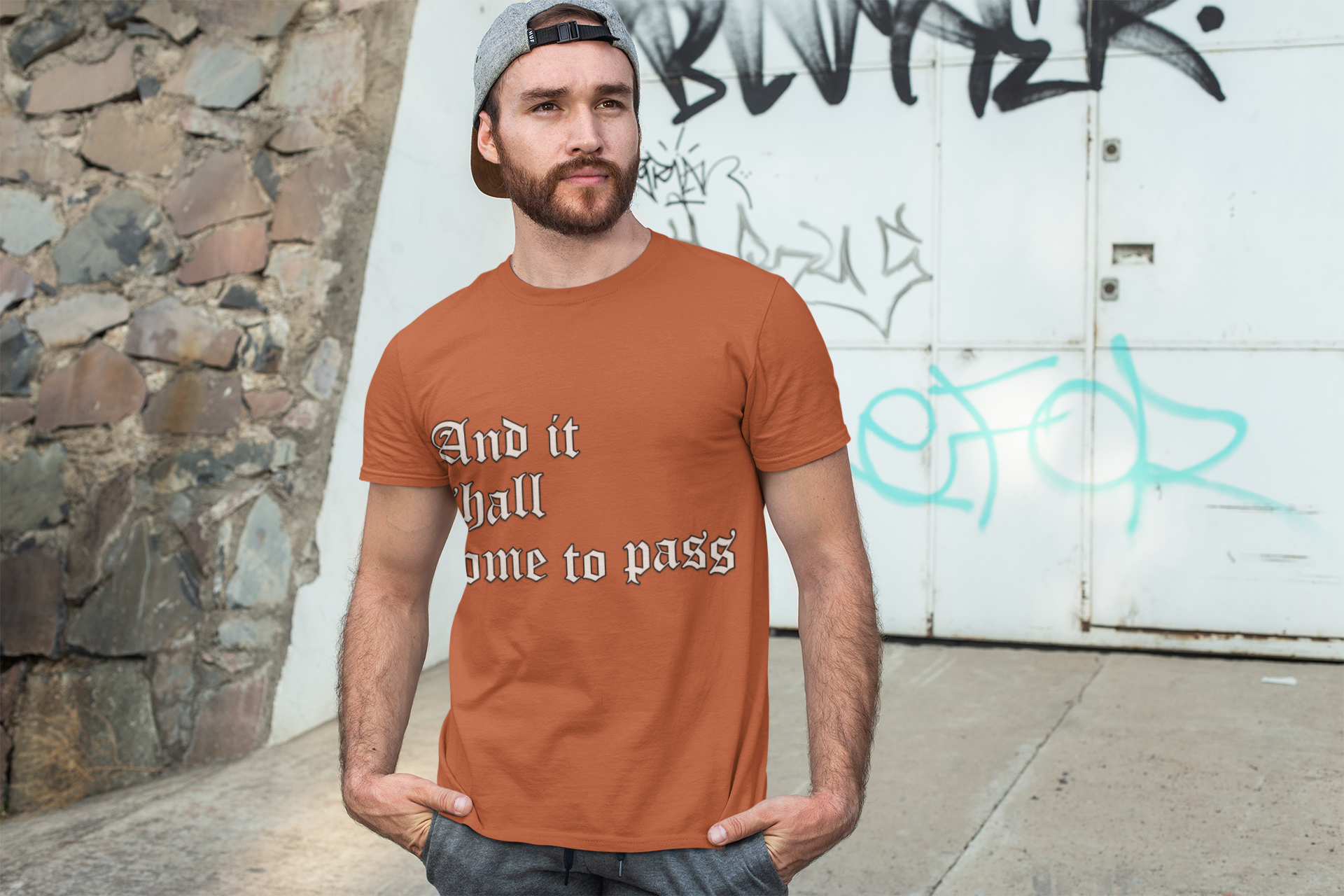 A man wearing an orange unisex short sleeve tee with a motivational quote, "And it shall come to pass" printed on the front.