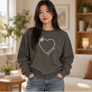 A woman wearing a dark gray crewneck sweatshirt with a white heart outline graphic on the front, standing in a cozy, softly-lit living room.